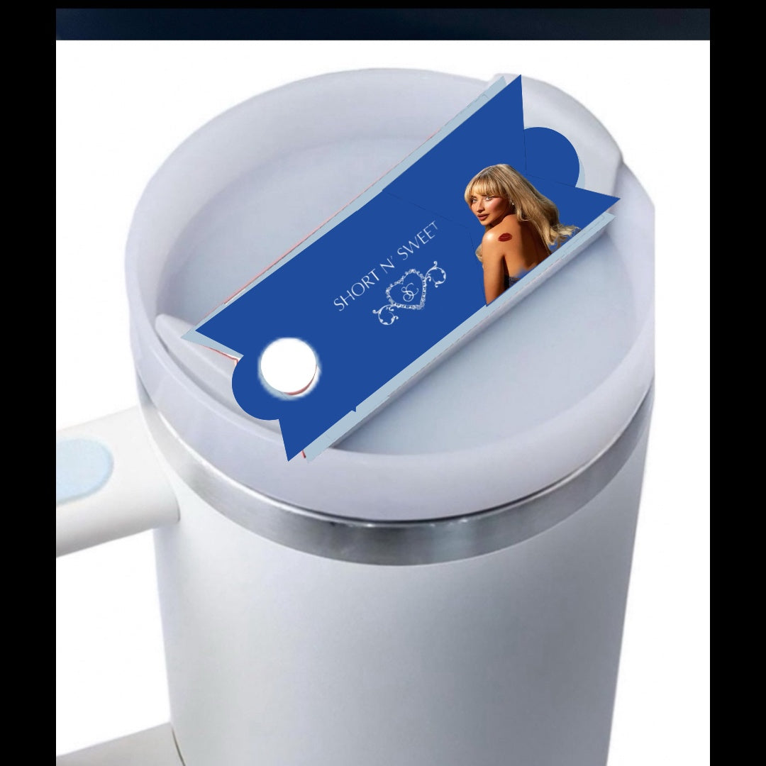 A white mug with a light blue button feature, covered by a lid. On the lid sits a blue, rectangular packaging featuring an image of a woman with blonde hair in a black outfit. The text on the packaging reads "SHOW 'N' SWEET." Crafted from premium materials, it resembles the Sabrina Carpenter Short & Sweet Stanley Lid Plate/Topper by Kreative Kreationz in quality.