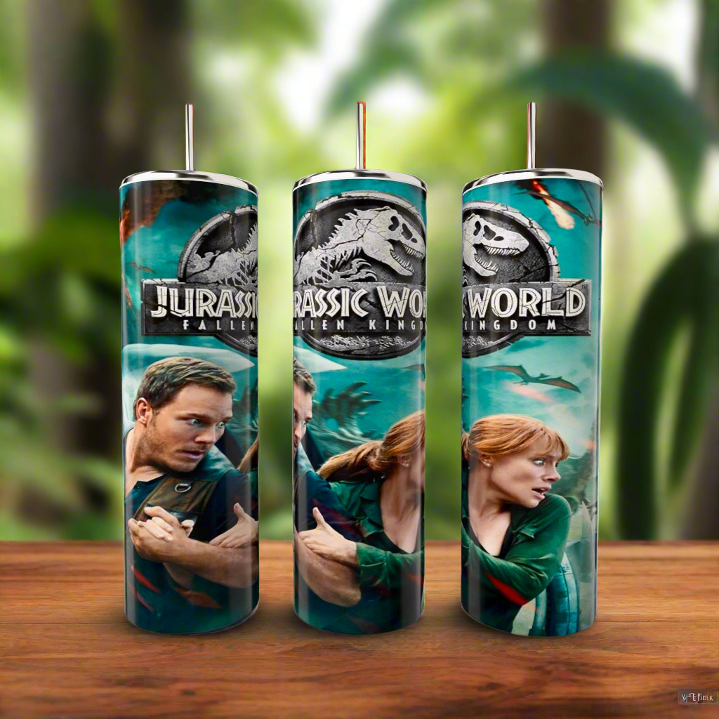 Three cylindrical candles with "Jurassic World: Fallen Kingdom" designs rest on a wooden surface. Nearby, a Kreative Kreationz Fallen Kingdom 20oz Tumbler, featuring double-wall insulation, stands against a blurred jungle background, enhancing the themed ambiance.