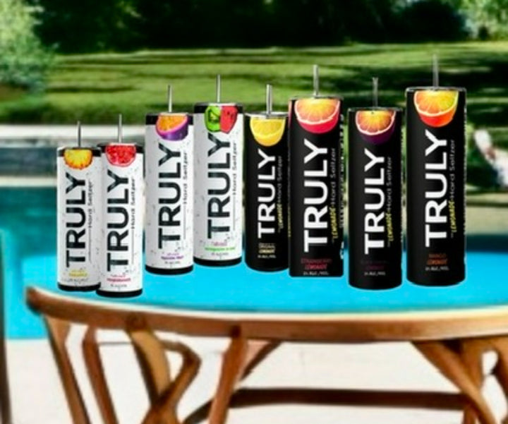 Cans of Truly hard seltzer in an array of flavors sit beside a Kreative Kreationz Truly Tumbler, featuring a 3D fruit and ice lid topper, with a scenic pool and lush greenery in the background.