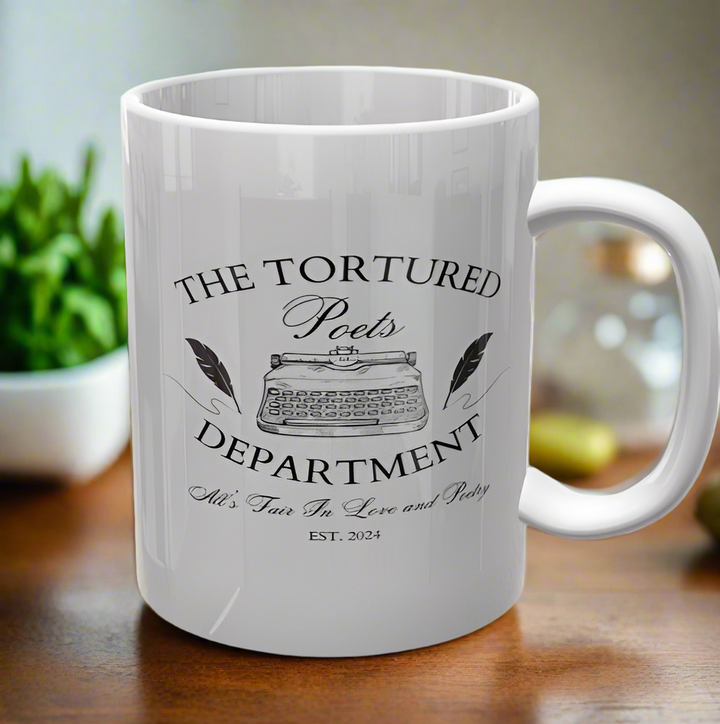 A white ceramic mug from Kreative Kreationz, named "The Tortured Poets Department | TTPD Ceramic Mug," features the text "The Tortured Poets Department" and an illustration of a vintage typewriter in the center. Below the illustration, it reads "All's Fair in Love and Poetry" and "Est. 2024." The mug is placed on a wooden surface with a plant in the background, making it perfect for any Taylor Swift fan.