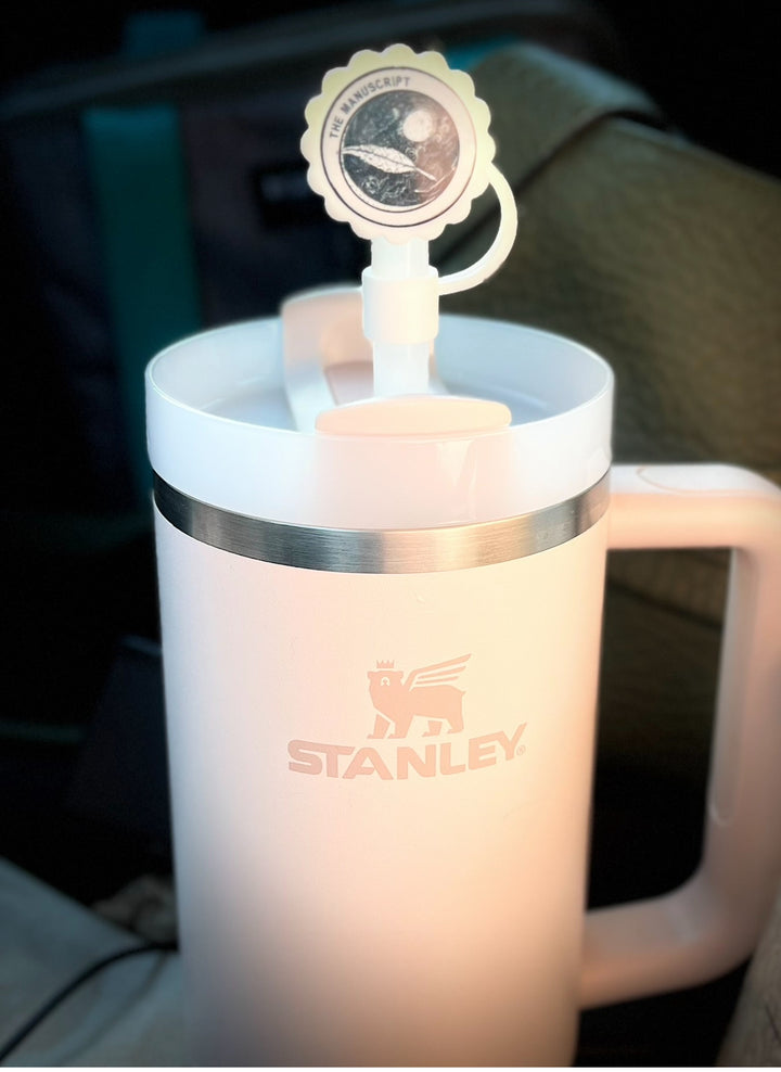 A white cup with a handle displays a round sticker on the lid, which features an illustration of a planet and spacecraft, accompanied by the text "THE MANUSCRIPT" around it. The background is black and the image appears in black and white. For added convenience, it pairs well with Kreative Kreationz's Straw Cover Topper, which fits Stanley cup sizes 40oz, 30oz, and 20oz with 10mm straws.