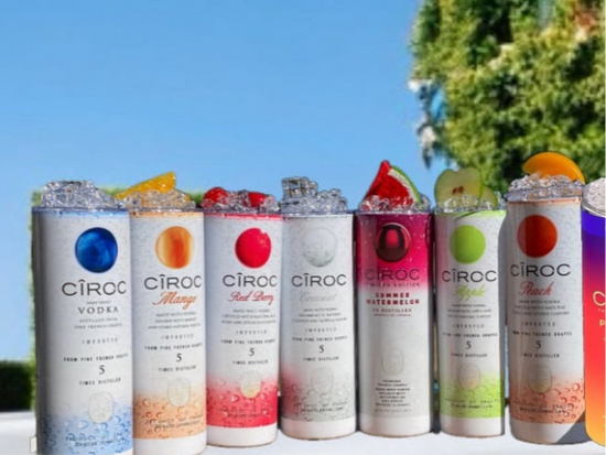 A lineup of CÎROC vodka cans in various flavors including original, mango, red berry, coconut, watermelon, and peach is beautifully arranged on an outdoor surface with a green hedge and blue sky background. Each can is paired with a chic 20oz Ciroc Tumbler with 3D Lid Topper by Kreative Kreationz.