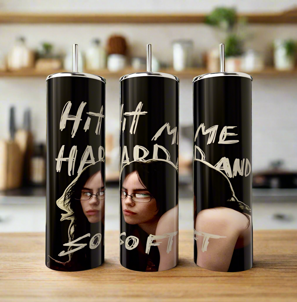 Three tall tumblers by Kreative Kreationz on a counter showcase dark designs of a woman with glasses, inspired by luxury. They read "Hit me hard and so soft" in bold text. The kitchen background is softly blurred. Product: Hit Me Hard And Soft Billie Eilish Tumbler.