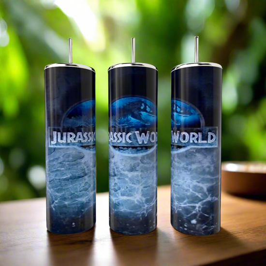 Three elegant Jurassic Park 20oz tumblers by Kreative Kreationz feature luxury-inspired stainless steel with silver lids and are showcased beautifully against ocean waves on a wooden surface, complemented by a blurred green foliage backdrop.