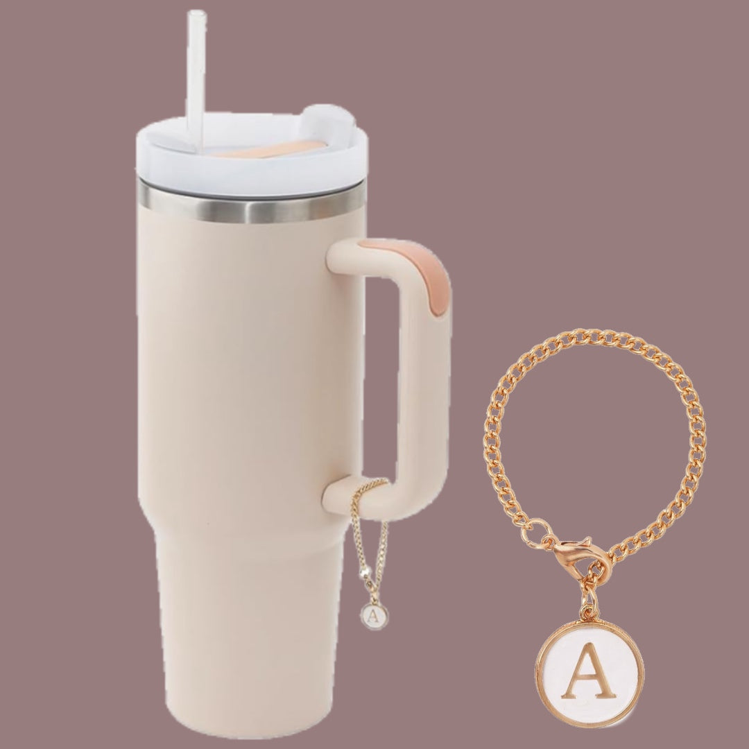 A beige insulated tumbler with a handle and straw from Kreative Kreationz is adorned with a Monogram Charm Dangle featuring the letter "A." It also comes with a matching gold chain bracelet, both showcasing the Monogram Charm Dangle with the letter "A." The background is solid taupe.