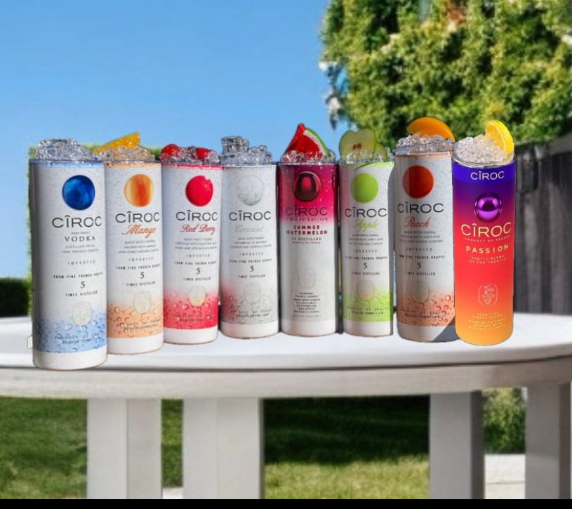 A lineup of CÎROC vodka cans in various flavors including original, mango, red berry, coconut, watermelon, and peach is beautifully arranged on an outdoor surface with a green hedge and blue sky background. Each can is paired with a chic 20oz Ciroc Tumbler with 3D Lid Topper by Kreative Kreationz.