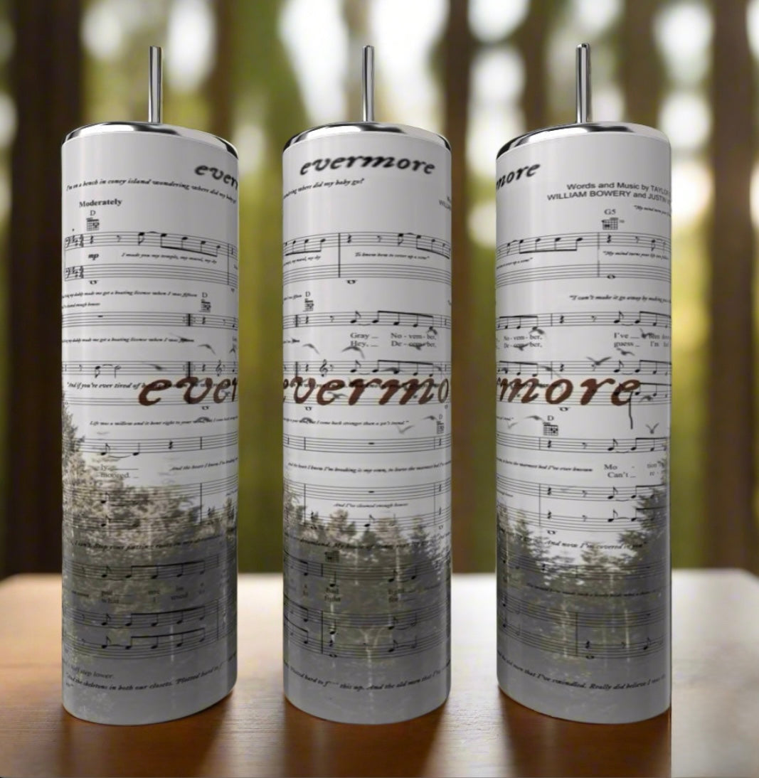 The Kreative Kreationz 20oz Skinny Tumbler features a printed sheet music design and the word "evermore," evoking the enchanting magic reminiscent of Taylor Swift's album, set against a wooden surface with a blurred forest backdrop.