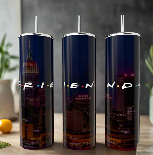 Three Kreative Kreationz "Friends City" 20oz tumblers with double-wall insulation showcase a nighttime city skyline and iconic "FRIENDS" lettering. They sit on a table with a blurred background featuring plants and an orange, making them perfect for fans of the beloved 90s sitcom!.