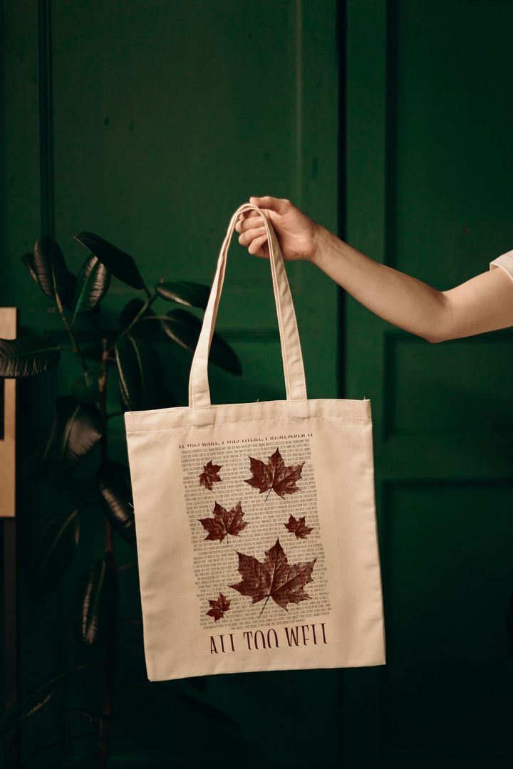 A person holds the Kreative Kreationz "All Too Well Tote Bag with lyrics," featuring scattered brown maple leaves on beige canvas. The phrase "All Too Well" appears at the bottom, with a blurred background of a green-leafed plant against a dark green wall.