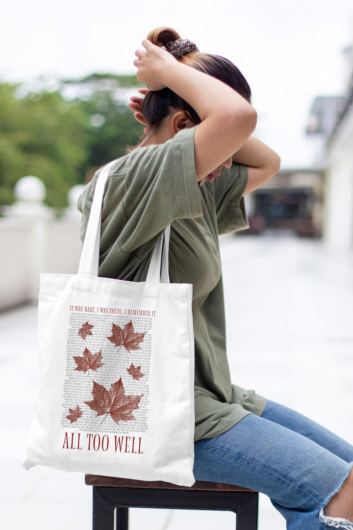 A person outdoors adjusts their hair in a green shirt and jeans, holding a white tote bag from Kreative Kreationz featuring autumn leaves and "ALL TOO WELL" lyrics, evoking Taylor Swift's storytelling, against a softly blurred greenery backdrop.