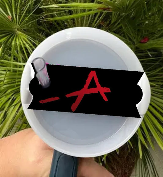 A person holds a white megaphone with a black sticker displaying a red, hand-drawn 'A', alluding to Pretty Little Liars. Behind it, green palm leaves enhance the intrigue. The sticker is the "Pretty Little Liars Stanley Lid Topper" by Kreative Kreationz.