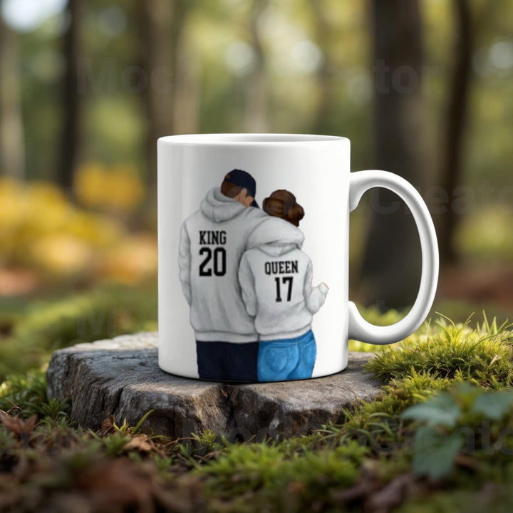 The Couples Ceramic Mug by Kreative Kreationz is a romantic high-quality gift featuring a couple in hoodies labeled "KING 20" and "QUEEN 17," standing with their backs together, artfully set on a tree stump in a serene forest.