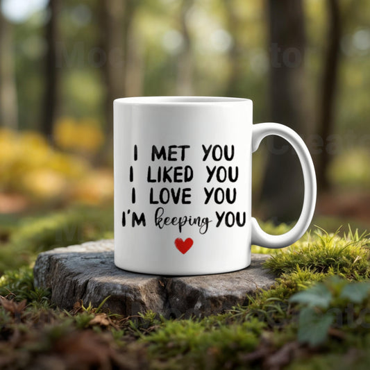 The Couples Ceramic Mug by Kreative Kreationz is made from high-quality ceramic and features black text "I met you, I liked you, I love you, I'm keeping you" with a red heart. Ideal as a romantic gift, it sits on a rock against a blurred forest backdrop.