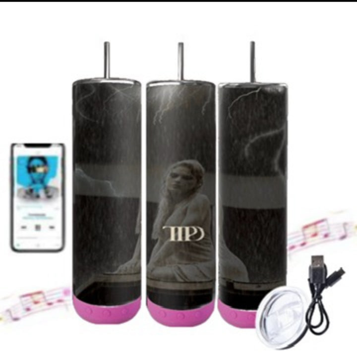 Three TTPD Bluetooth Tumblers by Kreative Kreationz, featuring a black cylindrical design with pink bases, are displayed. Each tumbler showcases a statue motif and the initials "MP." On the left, a smartphone screen displays a music app. A USB charging cable and a lid are placed beside the tumblers, with musical notes in the background.